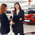 Cheerful young friendly dealer in formal stylish black dress showing contract to smiling female customer in black jacket while standing in car showroom against new shiny automobiles