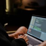 Focused hands typing code on a laptop in a dimly lit room, showcasing programming activities.