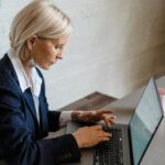 Professional woman in blazer working on laptop, seated at a desk with documents.
